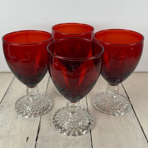 May include: Four vintage red wine glasses with clear, textured bases. The ruby-colored bowls of the glasses are set against a light-colored wooden surface. The glasses are likely made of glass.
