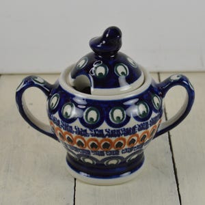 May include: A blue and white ceramic sugar bowl with a lid. The lid has a small bird finial. The bowl is decorated with a peacock feather pattern.