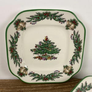 May include: Square ceramic Christmas plate with a green scalloped edge. The plate features a decorated Christmas tree in the center, surrounded by a garland of greenery, berries, and bells. The plate has a cream-colored background.