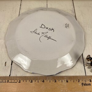 May include: A white ceramic plate with a scalloped edge, featuring the handwritten word "Dusk" and a signature. A small dog figurine is next to the plate. A wooden ruler is at the bottom.