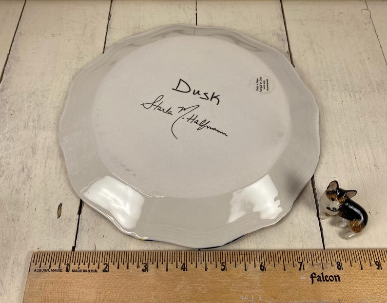 May include: A white ceramic plate with a scalloped edge, featuring the handwritten word "Dusk" and a signature. A small dog figurine is next to the plate. A wooden ruler is at the bottom.