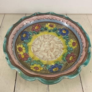 May include: A decorative ceramic bowl with a scalloped edge. The bowl features a central yellow design with a woven pattern, surrounded by colorful flowers in red, yellow, and blue. The rim has a pink and blue border, with a green edge.