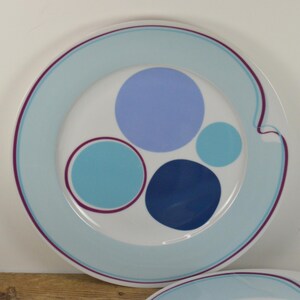 May include: A white ceramic plate with a blue and purple design. The plate has a blue rim and a swirl design in the center. The plate is decorated with four circles in different shades of blue.