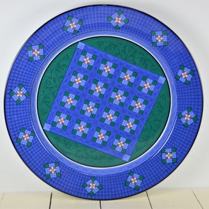 May include: A blue and green ceramic plate with a quilt pattern. The plate has a white border and a green center with a blue quilt pattern. The plate is decorated with small white flowers.