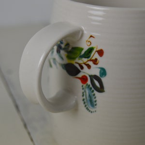 May include: White ceramic mug with a curved handle and floral design. The mug features a textured surface and is decorated with colorful hand-painted flowers, leaves, and berries in shades of blue, green, red, and yellow.