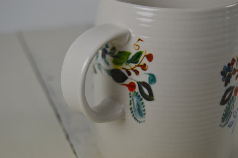 May include: White ceramic mug with a curved handle and floral design. The mug features a textured surface and is decorated with colorful hand-painted flowers, leaves, and berries in shades of blue, green, red, and yellow.