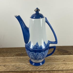 May include: A white and blue porcelain coffee pot with a matching cup. The coffee pot has a blue handle, spout, and lid. The cup is attached to the pot. The design features blue floral patterns. The coffee pot is on a wooden surface.