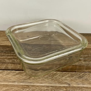 May include: A clear, square glass baking dish with a matching lid. The dish has rounded corners and a slightly raised lid. The glass is transparent, allowing visibility of the contents. The dish is sitting on a wooden surface.