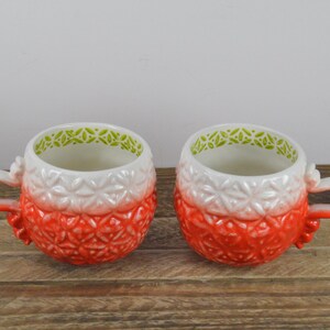 May include: Two white ceramic mugs with a red and white ombre design and green trim. The mugs have a textured surface and are perfect for hot beverages.