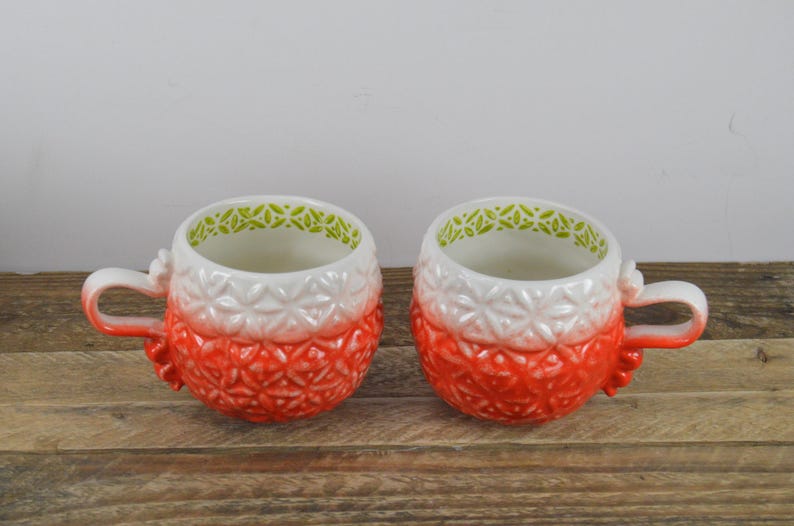 May include: Two white ceramic mugs with a red and white ombre design and green trim. The mugs have a textured surface and are perfect for hot beverages.
