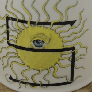 Pode incluir: Caneca de cer&acirc;mica branca com uma letra preta "S" e um sol amarelo com um olho azul. O sol tem raios amarelos.