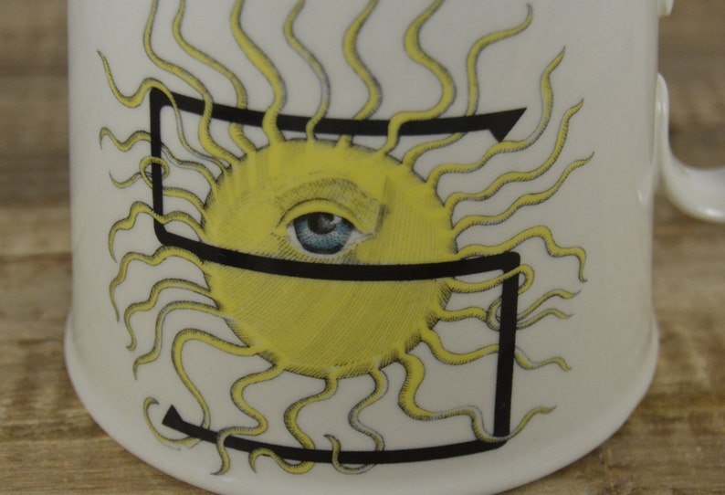 Pode incluir: Caneca de cer&acirc;mica branca com uma letra preta "S" e um sol amarelo com um olho azul. O sol tem raios amarelos.