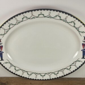May include: Oval serving platter with a white center, a black scalloped border, and a gold rim. The border features a green and white garland design. Two nutcracker figures in red, blue, and white are on the platter.