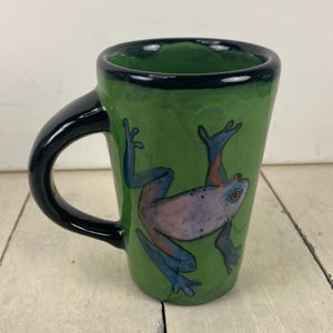 May include: Green ceramic mug with a black handle and rim. The mug features a hand-painted frog illustration in blue, pink, and orange. The mug is set against a white wooden surface.