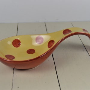 May include: A ceramic spoon rest with a yellow interior and a reddish-brown exterior. The spoon rest features a series of maroon and pink polka dots. The handle curves upward, providing a convenient place to rest a spoon while cooking.