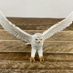 May include: A white owl figurine with outstretched wings, showcasing detailed feather textures and brown markings. The owl's eyes are focused forward, and its talons are visible. The figurine is set against a wooden background.