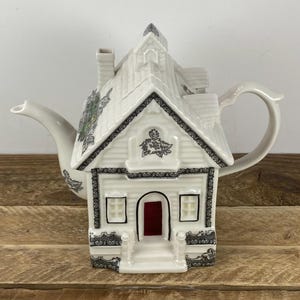 May include: A white ceramic teapot shaped like a house, featuring a red door and black trim. The roof has a chimney and is decorated with black floral patterns. The handle and spout are curved.