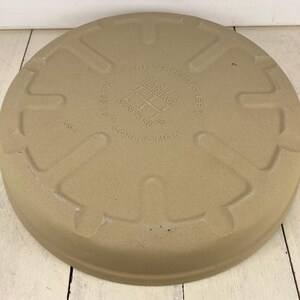 May include: A circular, tan stoneware pizza stone with embossed text reading "The Pampered Chef" and "Heritage Stoneware Collection". The stone has a raised edge and is made in the USA.