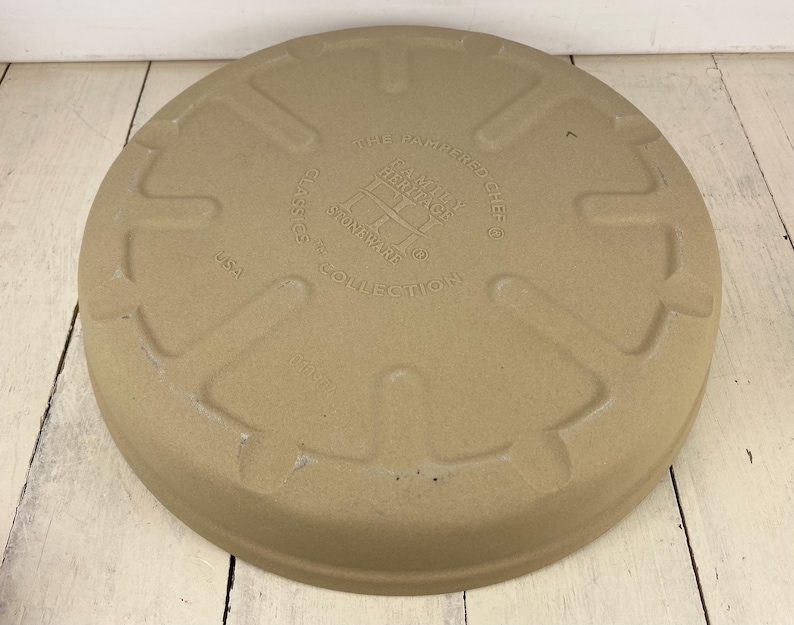 May include: A circular, tan stoneware pizza stone with embossed text reading "The Pampered Chef" and "Heritage Stoneware Collection". The stone has a raised edge and is made in the USA.