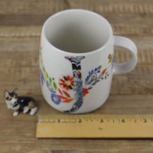 May include: White ceramic mug with a floral "J" design and a small corgi figurine. The mug has a handle and is decorated with colorful flowers and leaves. A wooden ruler is next to the mug. The corgi figurine is brown, black, and white.
