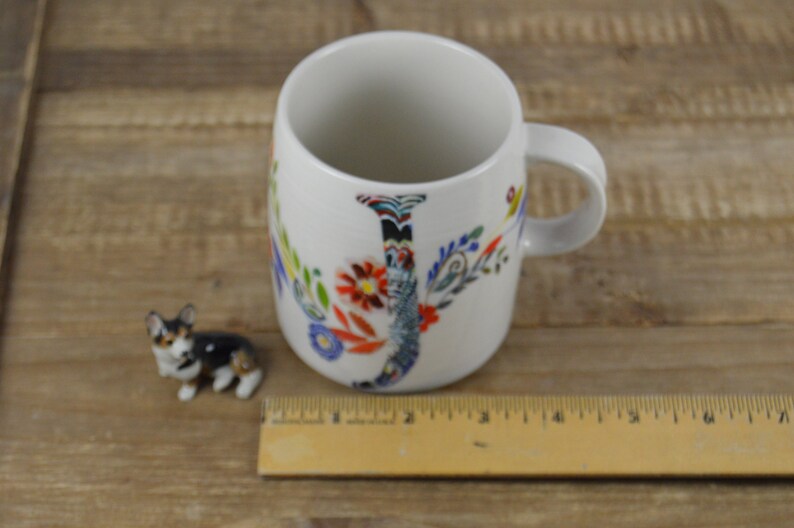 May include: White ceramic mug with a floral "J" design and a small corgi figurine. The mug has a handle and is decorated with colorful flowers and leaves. A wooden ruler is next to the mug. The corgi figurine is brown, black, and white.