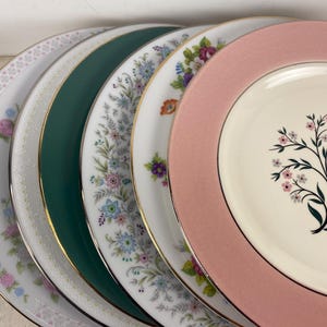 May include: A collection of vintage decorative plates with floral and geometric patterns. The plates feature a variety of colors, including pink, green, white, and gold. The plates are arranged in a semi-circle, showcasing their different designs.