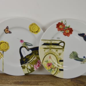 Puede incluir: Dos platos de cer&aacute;mica blanca con dise&ntilde;os florales y de p&aacute;jaros coloridos. Un plato presenta un colibr&iacute;, mariposas y flores amarillas. El otro plato presenta un p&aacute;jaro azul, flores rojas y un dise&ntilde;o dorado y negro.