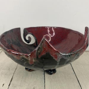 May include: A handmade ceramic bowl with a unique design. The bowl features a deep red interior and a black and gray exterior. It has a distinctive shape with a spiral cutout and three small feet. The bowl is suitable for decorative or functional use.