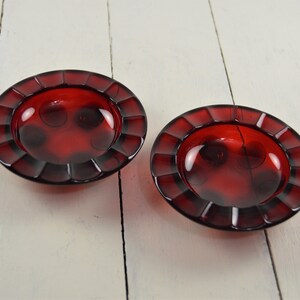 May include: Two red glass ashtrays with a fluted rim and a small indentation in the center.