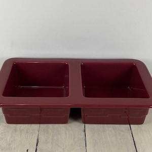 May include: A rectangular, burgundy-colored ceramic baking dish with two square compartments. The dish has a glossy finish and is set on a light-colored wooden surface. The dish is designed for baking.