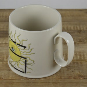 Pode incluir: Caneca de cer&acirc;mica branca com um sol amarelo com um olho azul e um desenho de linhas pretas.