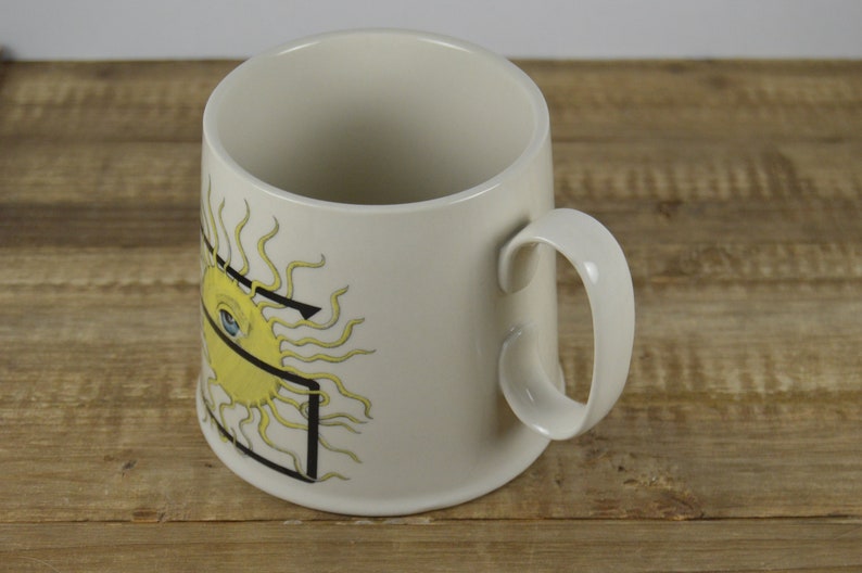 Pode incluir: Caneca de cer&acirc;mica branca com um sol amarelo com um olho azul e um desenho de linhas pretas.
