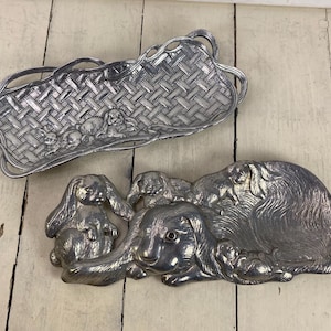 May include: Two silver-colored decorative trays. One is a rectangular basket weave design with bunny figures. The other is an oval-shaped tray with a large bunny and several smaller bunnies. The trays are on a white wooden surface.