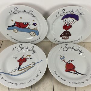 May include: Set of four white ceramic plates with whimsical Santa Claus illustrations. Each plate features a different scene with Santa, accompanied by the text "Santa Baby" and song lyrics. The plates have a playful, festive design.