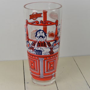 May include: A tall, clear glass with a red and blue illustration of a man with a mustache behind a bar. The glass has the text "Tram Yoodle" on it.