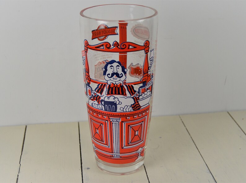 May include: A tall, clear glass with a red and blue illustration of a man with a mustache behind a bar. The glass has the text "Tram Yoodle" on it.