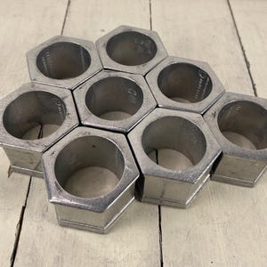 May include: A set of eight silver-colored, hexagonal napkin rings arranged in a honeycomb pattern. Each ring has a cylindrical opening and a geometric design. The napkin rings are made of metal and sit on a light-colored wooden surface.