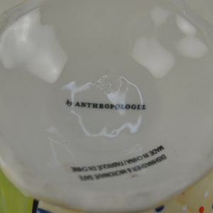 May include: White ceramic mug with a green and yellow rim and a colorful polka dot pattern. The mug has the text "by Anthropologie" and "Designed & Exclusive to Anthropologie Made in China" printed on the bottom.
