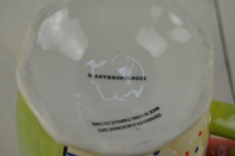May include: White ceramic mug with a green and yellow rim and a colorful polka dot pattern. The mug has the text "by Anthropologie" and "Designed & Exclusive to Anthropologie Made in China" printed on the bottom.