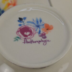 May include: A white ceramic bowl with a floral design and the text "Anthropologie" in purple. The bowl has a pink, blue, and orange floral pattern.