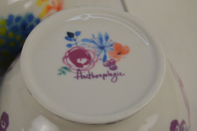 May include: A white ceramic bowl with a floral design and the text "Anthropologie" in purple. The bowl has a pink, blue, and orange floral pattern.