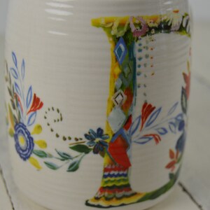 May include: White ceramic mug with a colorful floral design and the letter "J". The letter is decorated with a patchwork of patterns and colors, including blue, red, yellow, and green. The mug has a slightly textured surface.