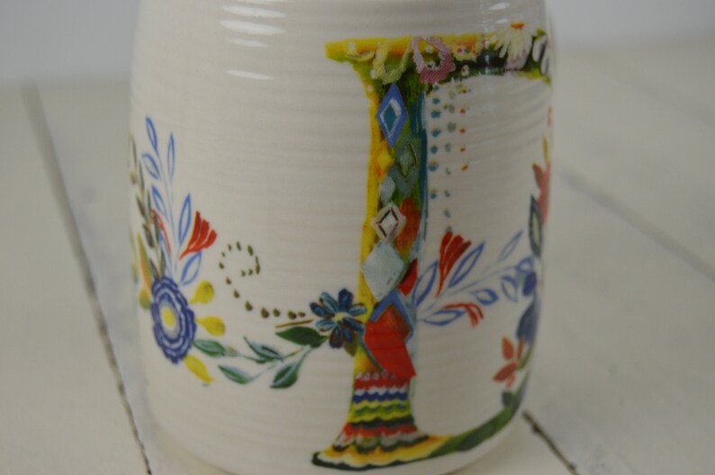 May include: White ceramic mug with a colorful floral design and the letter "J". The letter is decorated with a patchwork of patterns and colors, including blue, red, yellow, and green. The mug has a slightly textured surface.
