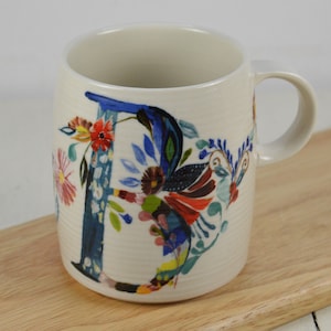 May include: White ceramic mug with a blue and multi-colored floral design featuring the letter "D".