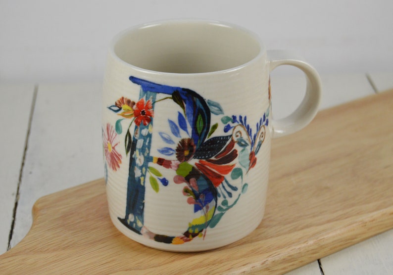 K&ouml;nnte beinhalten: Wei&szlig;e Keramiktasse mit einem blauen und mehrfarbigen Blumendesign mit dem Buchstaben "D".