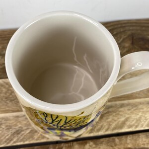 K&ouml;nnte beinhalten: Cremefarbene Keramik-Tasse mit rundem Henkel. Der Innenraum ist hellbeige, und die Au&szlig;enseite weist ein farbenfrohes, abstraktes Design mit gelben, blauen und braunen Farbt&ouml;nen auf. Die Tasse steht auf einer Holzoberfl&auml;che.