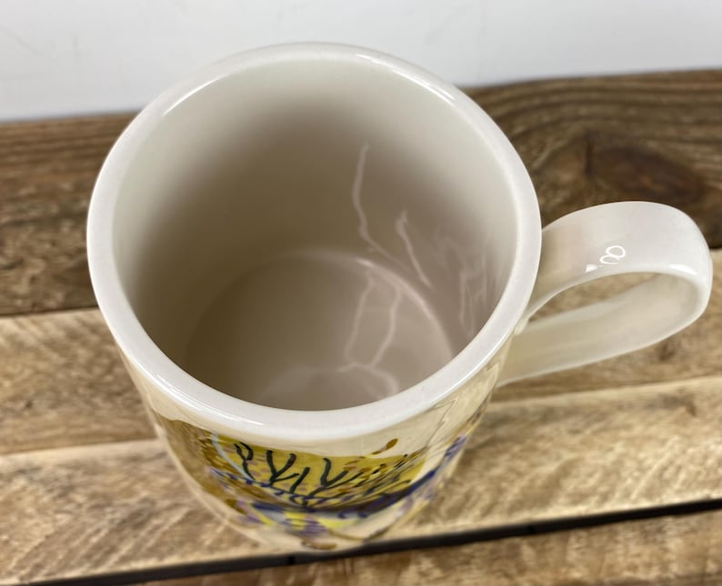 K&ouml;nnte beinhalten: Cremefarbene Keramik-Tasse mit rundem Henkel. Der Innenraum ist hellbeige, und die Au&szlig;enseite weist ein farbenfrohes, abstraktes Design mit gelben, blauen und braunen Farbt&ouml;nen auf. Die Tasse steht auf einer Holzoberfl&auml;che.