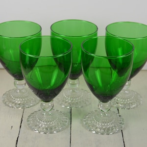 May include: Set of five green glass wine glasses with clear, textured bases. The glasses are arranged in a semi-circle on a white surface.