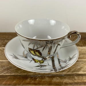 May include: White teacup and saucer set with a gold rim and handle. The design features silver and gold bamboo stalks and yellow butterflies. The teacup sits on a matching saucer, creating a cohesive and elegant look.