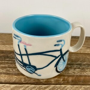 K&ouml;nnte beinhalten: Cremefarbene Keramiktasse mit blauem Innenraum und einem Fahrradmuster in Marineblau und Hellblau. Die Fahrradillustration umschlie&szlig;t die Tasse, mit rosa Akzenten am Lenker. Die Tasse hat einen gebogenen Griff.